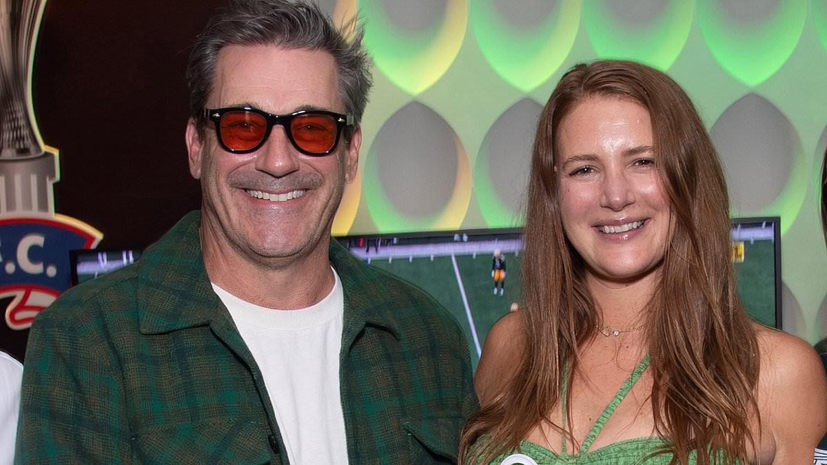 Jon Hamm, 55, and pregnant wife Anna Osceola, 38, pose with a New York Jets baby onesie as they head to Green Bay Packers game